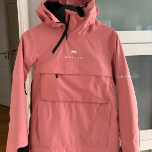 Rosa vindjacka från Montec med huva - Säljer en snygg rosa vindjacka från Montec med stor magficka, huva och svarta dragkedjor. Jackan har en clean look med logga framtill och på ärmen, samt justerbar huva. Den är så gott som ny då jag knappt använt den!