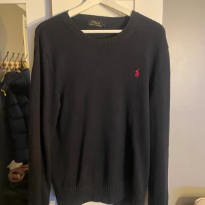 Svart stickad tröja Polo Ralph Lauren - Klassisk svart stickad tröja från Polo Ralph Lauren i 100% bomull. Tröjan har rund halsringning, långa ärmar och den ikoniska röda loggan broderad på bröstet. Perfekt för dig som gillar stilrena och tidlösa plagg.
