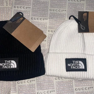 Svart och vit mössa The North Face - Två ribbstickade mössor från The North Face, en svart och en vit. Båda har uppvikt kant och tydlig logotyp framtill. Perfekt accessoar för kyliga dagar och enkel att matcha med olika outfits.