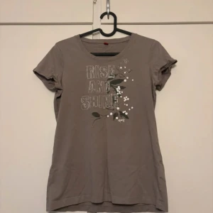 Brun t-shirt med tryck och blommor - Snygg brun t-shirt med texten 'Rise and Shine' framtill och blommigt tryck i vitt och grönt. Klassisk rund halsringning och korta ärmar. Perfekt för en avslappnad stil.