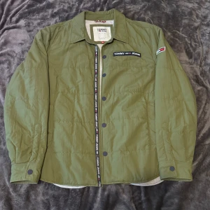 Olivgrön overshirt från Tommy Jeans XL - Snygg olivgrön overshirt från Tommy Jeans med tryckknappar och klassisk krage. Jackan har broderade loggor på bröstet och ärmen, samt detaljer med Tommy Jeans-band längs knäppningen. Materialet är lätt polyester och insidan är mörkblå. Perfekt för lager-på-lager.