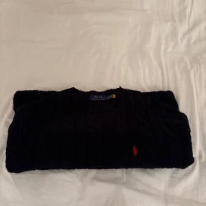 Mörkblå kabelstickad tröja Polo Ralph Lauren - Snygg mörkblå kabelstickad tröja från Polo Ralph Lauren med klassisk röd logobrodering på bröstet. Tröjan har rund hals och ribbade muddar. Perfekt för dig som gillar stilrena och tidlösa plagg.