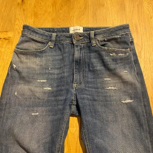 Dondup med slitningar  - Säljer ett par blå jeans från Dondup med snygga slitningar och klassisk femficksdesign. Jeansen har rak passform och normal midja, med diskreta detaljer och en cool tvättad look. Materialet är jeans i bomull och de har knappstängning framtill. Storlek 29
