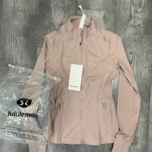 Ljusrosa träningsjacka från Lululemon - Snygg ljusrosa träningsjacka från Lululemon med dragkedja framtill och höga fickor med dragkedja. Jackan har en figurnära passform och hög krage, perfekt för aktiva dagar. Materialet är mjukt och stretchigt för maximal rörelsefrihet.