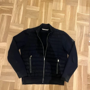Moncler Cardigan  - Snygg mörkblå cardigan från Moncler med quiltad front och två dragkedjeförsedda fickor. Cardiganen har ribbad krage och Moncler-logga på ärmen. Perfekt för dig som gillar stilren design och exklusiva märken.