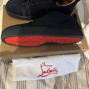 Mörkblå sneakers från Christian Louboutin - Snygga mörkblå sneakers från Christian Louboutin i mocka med klassisk röd sula. Skorna har svarta snören, rund tå och platt sula. Insidan är ljusbeige och de levereras med originalkartong och dustbag. Perfekta för dig som vill ha en lyxig och stilren look. I storlek 40 pris går att diskutera 