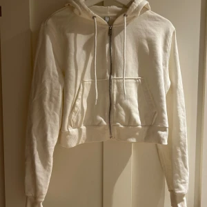 Vit croppad hoodie från H&M - Säljer en vit croppad hoodie från H&M i storlek L. Tröjan har huva med snörning, dragkedja framtill och två fickor. Materialet är mjuk bomull. Perfekt för chill dagar och enkel att matcha med jeans eller leggings.