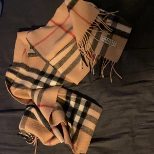 Beige rutig halsduk från Burberry - äkta klassisk beige halsduk från Burberry med det ikoniska rutiga mönstret i svart, vitt och rött. Halsduken är tillverkad i en lyxig mix av 50% kashmir och 50% ull.