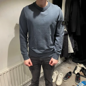 Stone island tröja - Snygg blå sweatshirt från Stone Island med klassisk rund hals och ribbade muddar. Tröjan har den ikoniska Stone Island-loggan på vänster ärm, vilket ger en extra cool detalj. Perfekt för dig som gillar streetwear och vill ha något stilrent i garderoben.