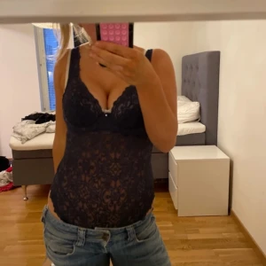 Svart spetsig korsettlinne - Säljer ett svart korsettlinne i genombruten spets med djup v-ringning och breda axelband. Toppen har en feminin, figurnära passform och en liten dekorativ detalj framtill. Perfekt för att lyfta vilken outfit som helst. Kan ej tyga lappen vart den är ifrån och storlek men passar S/M