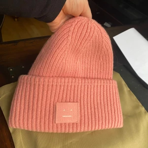 Rosa ribbstickad mössa Acne Studios - Pudder rosa /gammal rosa färg , ribbstickad mössa från Acne Studios med ikonisk face-patch framtill. Tillverkad i 100% ull för extra värme och mjuk känsla. Perfekt för att lyfta din vinterstil med en poppig färg och trendig look.        Storlek 05/56 cm Använd några gånger syns lite på insida att det var använd . Handtvätt rekommenderas men jag har inte provad tvätta.