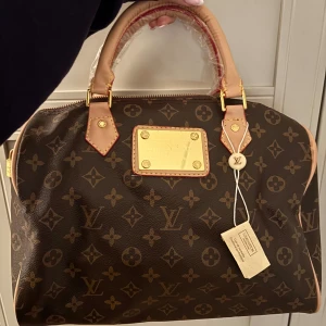 handväska - Snygg handväska med klassiskt monogrammönster i mörkbrunt och beige. Väskan har guldfärgade detaljer, dragkedja och två handtag med ljusbruna läderdetaljer. Insidan har fack och en avtagbar axelrem. Perfekt för dig som gillar lyxig design.