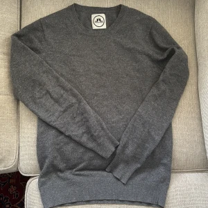 Grå kashmirtröja från J.Lindeberg - Mjuk och stilren grå tröja i 100% kashmir från The Bridge Project från J.lindeberg. Klassisk rund halsringning, ribbade muddar och lång ärm. Perfekt för dig som gillar en clean och tidlös look med premiumkänsla. Litet hål på armen, därav priset. Nypris runt 2000kr