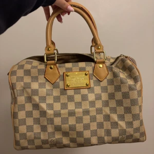 rutig handväska i skinn - Snygg handväska med klassiskt rutigt mönster i beige och grått. Väskan har detaljer i ljust skinn, guldfärgade metalldelar och dragkedja upptill. Perfekt storlek för det viktigaste och har ett innerfack med dragkedja.