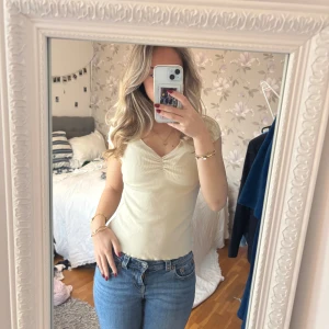 Ljusbeige ribbad v-ringad topp - Säljer en ljusbeige ribbad topp med v-ringning och lätt rynk framtill. Toppen är ärmlös och har en slim passform som sitter snyggt till jeans eller kjol. Perfekt för dig som gillar en enkel men stilren look.