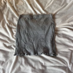 Grå ribbad kortkjol - Säljer en grå ribbad kortkjol med vågig kant nertill. Kjolen har en slim passform och är tillverkad i mjukt, stretchigt material som sitter skönt. Perfekt för dig som gillar enkel och stilren design med lite edge.