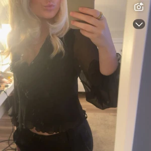 Svart croppad blus med volang - Svart, transparent och croppad blus med v-ringning och långa, luftiga ärmar. Blusen har volangdetaljer vid ärmslut och nederkant för en extra snygg touch. Perfekt för dig som gillar en trendig och luftig stil. Det är storlek m men sitter mer som en s😍