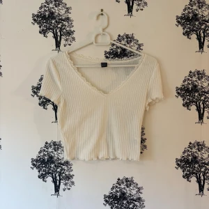 Vit ribbad croppad topp från SHEIN - Säljer en vit croppad topp från SHEIN i ribbad bomullsmix. Toppen har korta ärmar, v-ringning med spetskant och en mjuk, feminin look. Perfekt för dig som gillar enkla men söta detaljer. Passar dig som vill ha en slim fit och gillar ljusa färger.