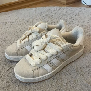 Beige adidas campus 00s sneakers i mocka - Populära adidas campus sneakers med klassiska vita ränder på sidorna. Skorna har breda beiga snören och en platt gummisula i ljusbrunt. Ovandelen är i mjuk mocka. Väldigt bekväma. Endast använda 3-4 gånger. Finns hund i hemmet. Storlek 38 2/3.