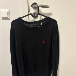 Svart tröja från Polo Ralph Lauren - Svart långärmad tröja från Polo Ralph Lauren i storlek M. Klassisk rund hals och röd broderad logga på bröstet. Tröjan är stickad och har ribbade muddar vid ärmslut och nederkant. Perfekt för dig som gillar stilrena och tidlösa plagg.