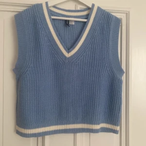 Blå stickad väst från H&M Divided - Snygg ljusblå stickad väst från H&M Divided med v-ringning och vita ränder vid halsringning och nederkant. Västen har en klassisk, ribbstickad struktur och är perfekt att styla över en skjorta eller t-shirt.