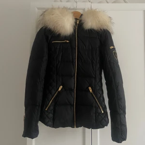 Svart dunjacka Rockandblue med äkta dun och päls - Snygg svart dunjacka i modell ”rita” från Rockandblue. Avtagbar luva och avtagbar pälskrage. Äkta päls (tvättbjörn) Jackan är figursydd och har guldiga dragkedjor. Perfekt för kalla dagar. Använd en vinter, pälsen är väl omtagen.