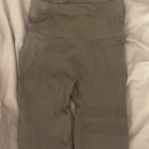 Beige ribbade leggings från Stronger - Snygga beige leggings från Stronger i storlek XS. De har ribbad struktur och bred hög midja som sitter skönt och ger extra stöd. Perfekta för träning eller chill, med stilren design och diskret logga upptill. Materialet är mjukt och stretchigt syntet.