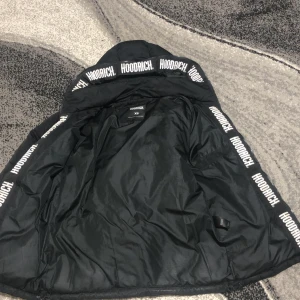 Svart pufferjacka från Hoodrich XS - Svart pufferjacka från Hoodrich i storlek XS. Jackan har huva med Hoodrich-logga runt kanten, dragkedja framtill och två sidofickor med dragkedja. Puffad design och långärmad modell, perfekt för kalla dagar. Snygg streetwear-vibe med tydlig branding.