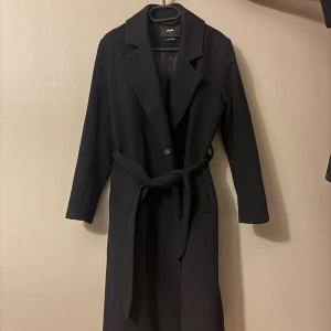 Svart kappa från Bikbok med bälte - Svart lång kappa från Bikbok i ullmix, med klassisk krage och knytbälte i midjan. Stilren och enkel design med raka linjer och långa ärmar. 