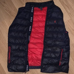 Polo Ralph Lauren Väst - Snygg dunväst från Polo Ralph Lauren i svart med röd insida och detaljer. Västen har quiltad design, ståkrage och dragkedjor på fickorna. Märkets logga broderad på bröstet och Polo-tryck längs insidan. Perfekt på våren samt hösten.