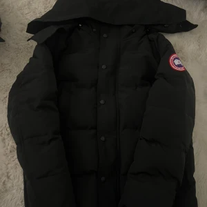 Svart dunjacka från Canada Goose - Svart dunjacka från Canada Goose med klassisk patch på ärmen. Jackan har huva, knappar framtill och ett puffigt quiltat utförande. Perfekt för kalla dagar och har en clean, stilren look som funkar till det mesta.