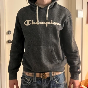 Mörkgrå Champion hoodie i medium - Snygg mörkgrå hoodie från Champion i storlek medium. Tröjan har vit Champion-logga på bröstet, vita snören i huvan och en klassisk känguruficka framtill. Perfekt för vintern.