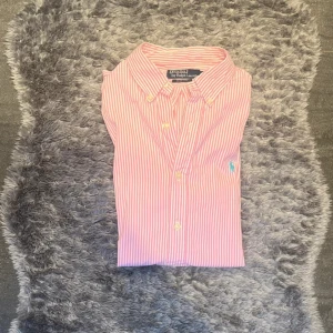 Rosa randig skjorta från Polo Ralph Lauren - Snygg rosa och vit randig skjorta från Polo Ralph Lauren i slim fit-modell. Skjortan har klassisk krage med knappar, lång ärm och det ikoniska blå broderade Polo-logot på bröstet. Tillverkad i mjuk bomull som känns skön mot huden.