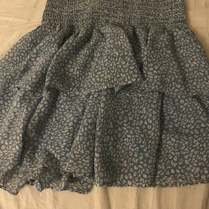 Blå leopardmönstrad volangkjol Chelsea - Supersöt blå kortkjol från Chelsea med ljusa leopardfläckar och volanger. Kjolen har smockad midja som sitter skönt och ger en snygg passform. Perfekt för dig som gillar djurmönster och vill ha en kjol med lite extra volym.