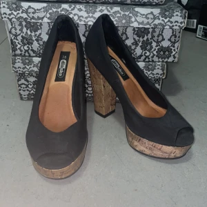 Svarta pumps med korkklack från Din Sko - Snygga svarta pumps från Din Sko med öppen tå och rejäl plattform samt hög klack i kork-look. Skorna har en klassisk rundad form och är perfekta för dig som vill sticka ut med en cool detalj på klacken. Materialet är textil och insidan är ljusbrun.