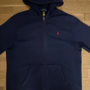 Marinblå hoodie från Polo Ralph Lauren - Snygg navyblue hoodie från Polo Ralph Lauren med dragkedja och huva. Tröjan har det klassiska röda Polo-logot på bröstet, känguruficka framtill och långa ärmar. Perfekt för chill dagar och enkel att matcha med jeans passform den passar mig som är 173cm lång