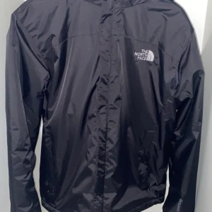 Svart vindjacka The North Face S - Svart vindjacka från The North Face Summit Series i storlek S Jackan har huva, dragkedja framtill med logga, och två sidofickor. Materialet är lätt och slitstarkt, perfekt för blåsiga dagar. Insidan har detaljer med The North Face-band och etikett.