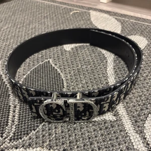 Svart och vit Dior bälte med CD-spänne - Snyggt bälte från Dior med ikoniskt CD-spänne i silver. Bältet har ett svart och vitt mönster med Dior-logga längs hela bandet. Tillverkat i skinn med textil på ovansidan och slätt svart läder på insidan. Perfekt accessoar för att lyfta din outfit.