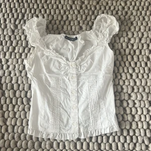 Vit blus med volang från Brandy Melville - Supersöt vit blus från Brandy Melville med volangdetaljer vid axlar och nederkant. Blusen har knappar framtill, rynkade axlar och fina veckade detaljer på framsidan. Perfekt för dig som gillar romantisk och feminin stil. Stl XS/S