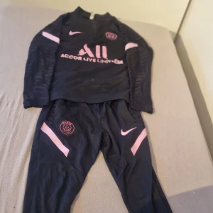 Svart PSG Nike träningströja med rosa detaljer - Svart långärmad träningströja från Nike x PSG med rosa detaljer och klubbmärke på bröstet. Tröjan har halv dragkedja vid halsen och är perfekt för fotbollsträning. Snygg sportig look med rosa Nike-logga och Paris Saint-Germain-tryck.