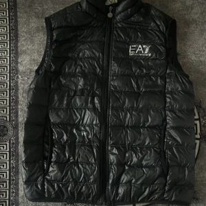 Svart dunväst från Emporio Armani EA7 - Stilren svart dunväst från Emporio Armani EA7 med quiltad design och dragkedja framtill. Västen har logga på bröstet och hög krage. Perfekt för lager-på-lager och ger en sportig vibe till din outfit.
