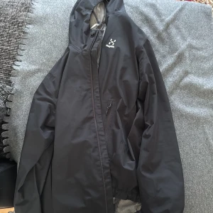 Svart vindjacka från Haglöfs - Svart vindjacka från Haglöfs med huva och meshfoder. Jackan har en snygg minimalistisk design, dragkedja framtill och en praktisk ficka med dragkedja på bröstet. Perfekt för dig som gillar att vara ute och vill ha en lätt jacka med cool look.