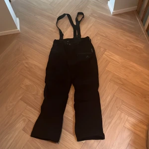 Svarta täckbyxor med hängslen - Mc Kinley svarta täckbyxor med justerbara hängslen och dragkedja fram. Byxorna har raka ben och praktiska fickor med dragkedja på sidan. Perfekta för kalla dagar och vinteraktiviteter. Storlek 152