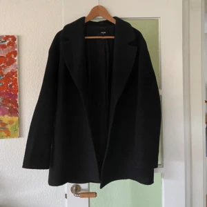 Svart kappa från Bik Bok i ullmix - Svart kappa från Bik Bok i storlek M, tillverkad i ullblandning. Klassisk rak modell med krage och två knappar framtill. Långärmad och med snyggt fall, perfekt för dig som gillar stilrena och enkla plagg.