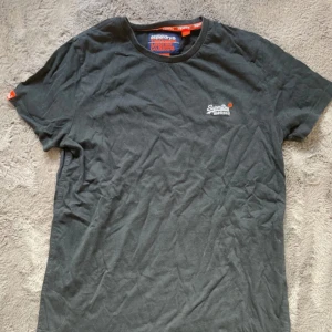 Svart Superdry t-shirt  - Svart t-shirt från Superdry med broderad logga på bröstet och orange detaljer vid hals och ärmslut. Klassisk rund hals och korta ärmar, perfekt för en chill och stilren look.