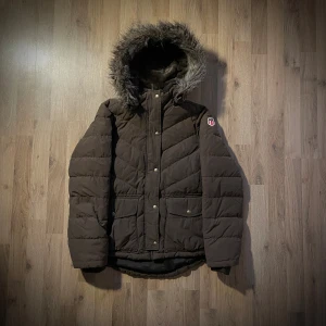 Brun quiltad dunjacka Polo Ralph Lauren M - Snygg brun dunjacka från Polo Jeans Company med fluffig fuskpäls på huvan och broderad RL-patch på ärmen. Jackan har quiltad design, tryckknappar, två stora fickor och långärmad passform. Perfekt för kalla dagar med sin varma och stilrena look.