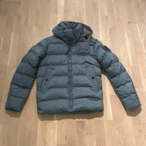 Grå pufferjacka från G-Star RAW - Snygg grå pufferjacka från G-Star RAW med huva och dragkedja framtill. Jackan har två snedställda fickor med knappar och en extra detaljficka vid bröstet. Perfekt för kalla dagar med sin vadderade design och stilrena look.