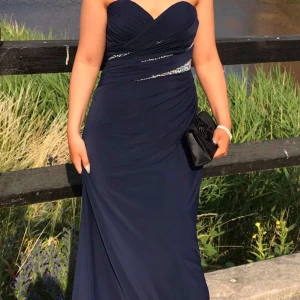 Marinblå långklänning med krisralldetalj - Elegant marinblå långklänning med hjärtformad överdel och draperad design. Klänningen är axelbandslös och har glittrande kristaller i midjan som ger extra lyxig känsla. Perfekt för bal eller festligare tillfällen. Använd vid 1 tillfälle. Storlek XS passar även de som pendlar mellan S och XS, köpt i en balk och bröllopsklänningsbutik. Nypris: 2999kr. Pris kan alltid diskuteras! Fler bilder? Skicka ett meddelande☺️