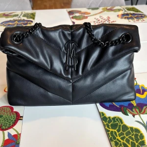 Svart quiltad axelväska Steve Madden - Snygg svart axelväska från Steve Madden med quiltad design och kedjerem i svart metall. Väskan har ett stilrent SM-emblem framtill och praktiskt innerfack med dragkedja. Perfekt storlek för det viktigaste och ger en cool vibe till din outfit. Aldrig använd kvitto finns. Kan skicka bild på det om det behövs.