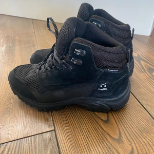 Svarta kängor från Haglöfs - Svarta kängor från Haglöfs med robust sula och snörning. Ovandelen är i mesh och mocka för extra andningsförmåga och slitstyrka. Perfekta för utomhusaktiviteter och äventyr. Märket syns tydligt på sidan och insidan av skon. Storlek 38 1/3